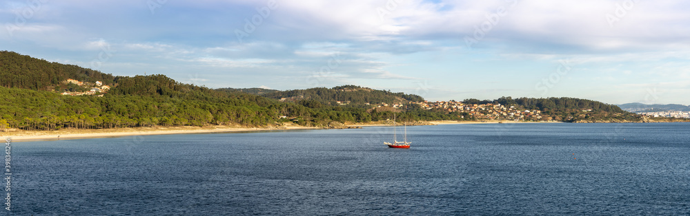 panorama view of a secluded golden sand beach with forest behind and a sailboat at anchor in the bay
