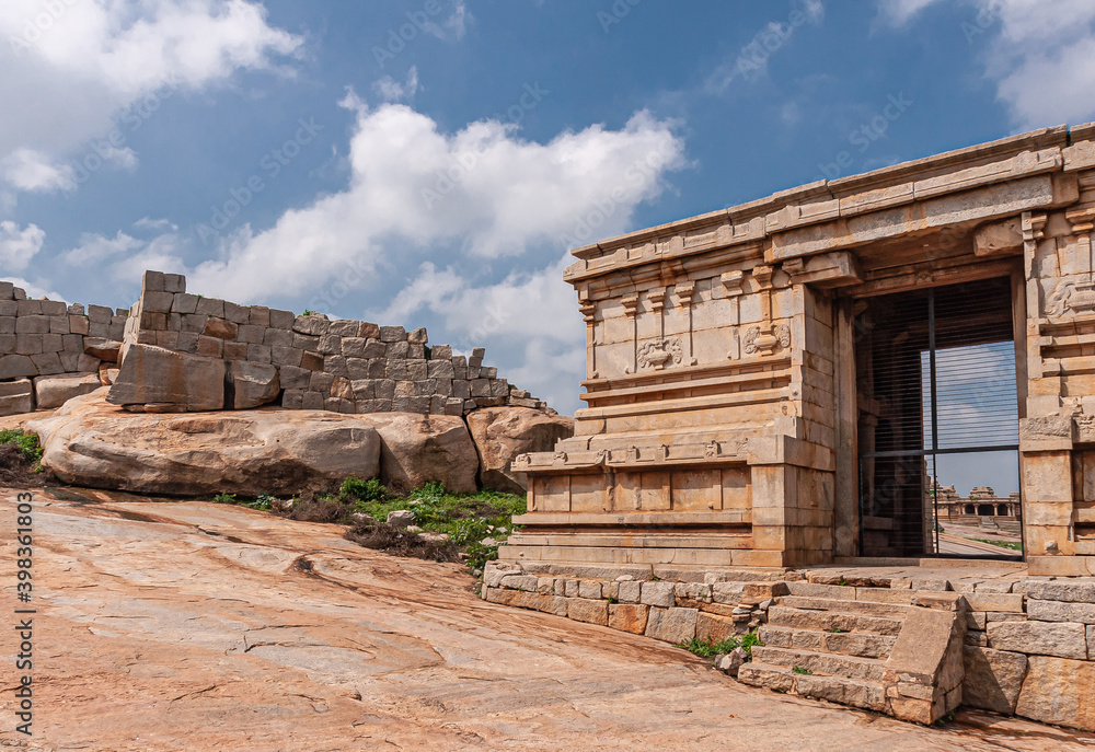 Hampi, Karnataka, India - November 4, 2013: Ruinous Kadelekalu Ganesha temple. Side brown stone buildings with screen and look-through. Gray stone wall and blue cloudscape.