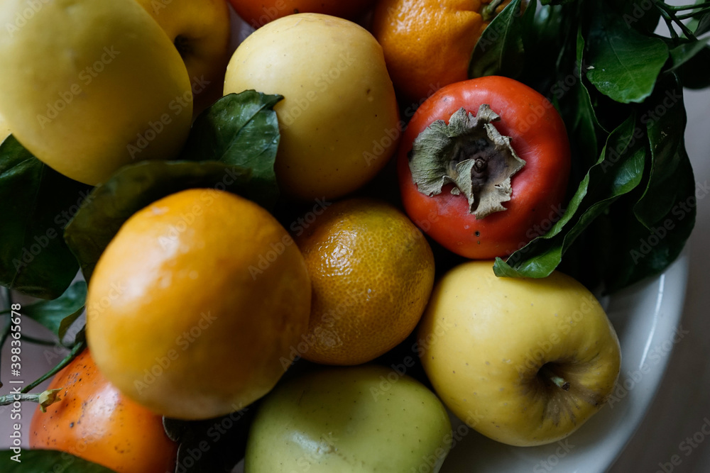 Brightly colored, orange and yellow, winter fruit: persimmon, apples, lemons and oranges arranged on a platter. Some green leaves. 