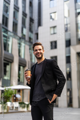 Coffee break. Confident young man in full suit holding coffee cup and looking away while standing outdoors with cityscape