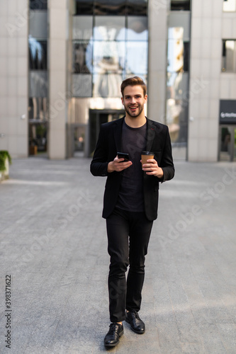 Work and coffee break. Cheerful young businessman using smartphone and holding cup of beverage.