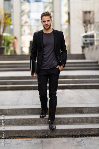 Handsome young businessman holding his laptop while using his phone and standing on the street