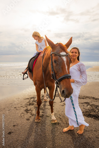 Horse riding on the beach. Cute little girl on a brown horse. Her mom standing near by. Love to animals. Mother and daughter spending time together. Selected focus.