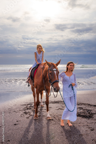 Horse riding on the beach. Cute little girl on a brown horse. Her mom wlking near by. Love to animals. Mother and daughter spending time together. Selected focus.