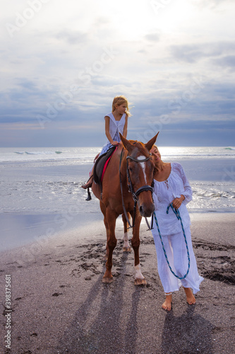 Horse riding on the beach. Cute little girl on a brown horse. Her mom standing near by Her mom standing near byv. Love to animals. Mother and daughter spending time together. Selected focus.
