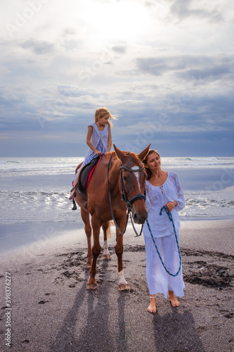 Horse riding on the beach. Cute little girl on a brown horse. Her mom standing near by. Love to animals. Mother and daughter spending time together. Selected focus.