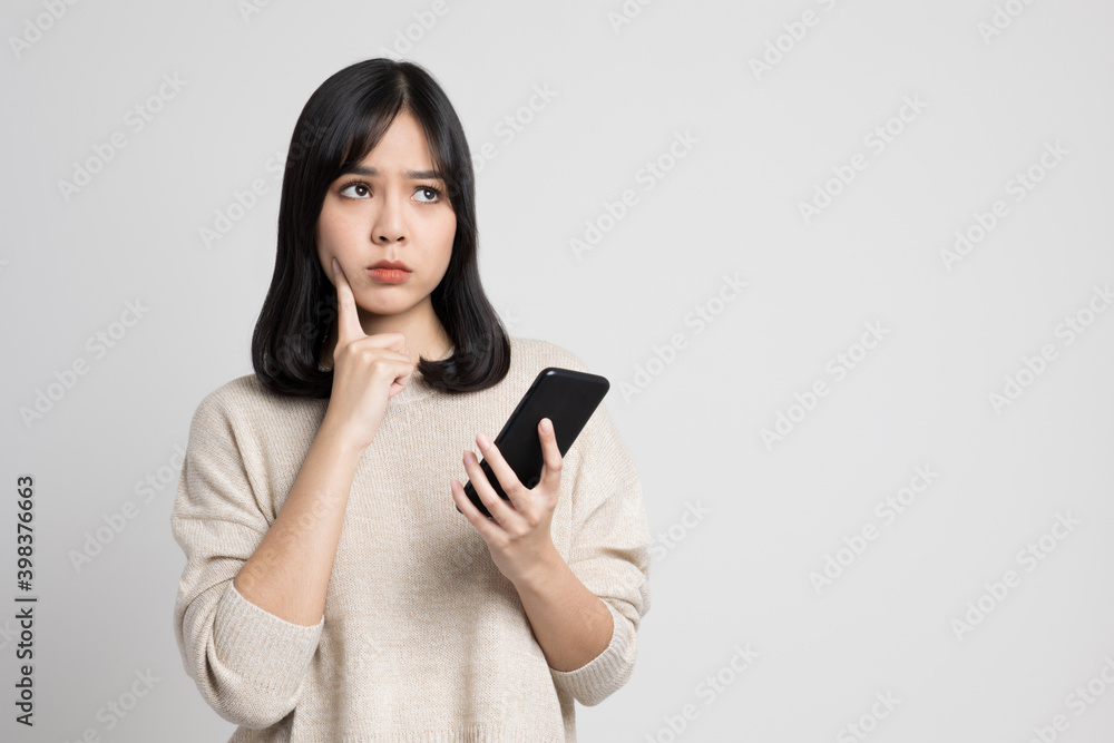 Beautiful young asian woman holding smartphone standing on isolated white background. She is looking at blank space and thinking something. Beautiful young asian woman holding smartphone standing on isolated white background. She is looking at blank space and thinking something.