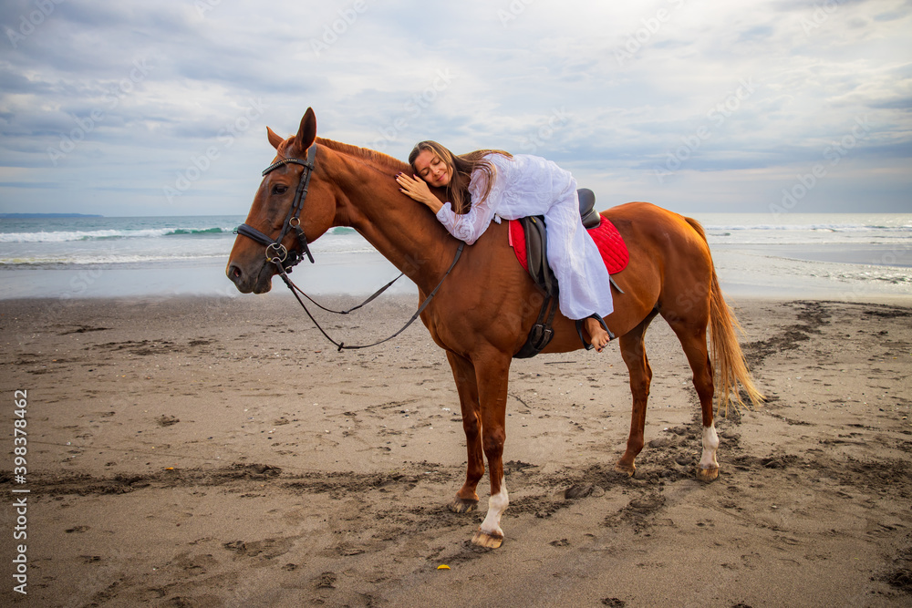Horsewoman lays down on the withers of the horse. Caucasian woman in white dress riding horse on the beach. Copy space. Sunset time on the beach. Outdoor activities.