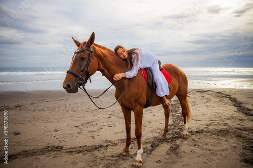 Horsewoman lays down on the withers of the horse. Caucasian woman in white dress riding horse on the beach. Copy space. Sunset time on the beach. Outdoor activities.