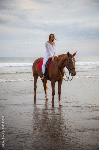 Beautiful caucasian woman riding horse by the sea. Female wearing white clothes. Copy space. Sunset time on the beach. Outdoor activities. © Olga
