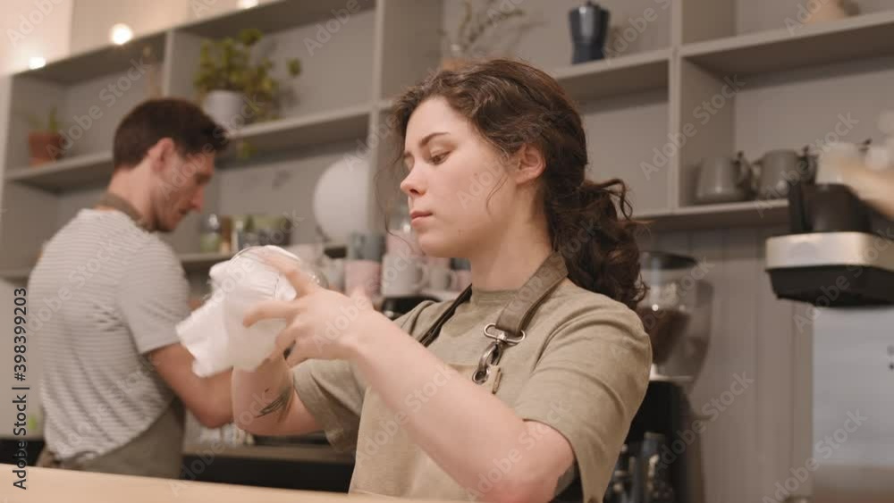Close-up POV of young Caucasian woman standing in bar, wiping glass with clean white cloth, looking through it at light, then smiling on camera. Male colleague working behind her Close-up POV of young Caucasian woman standing in bar, wiping glass with clean white cloth, looking through it at light, then smiling on camera. Male colleague working behind her