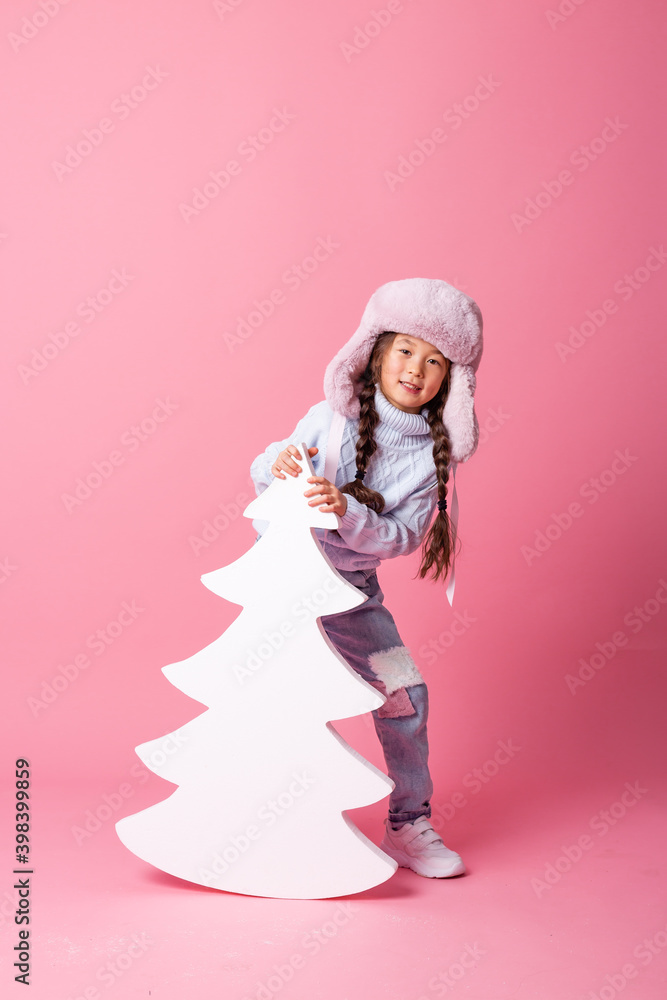 A cute little Asian girl in a winter hat stands next to a Christmas tree on a pink background. winter concept, space for text A cute little Asian girl in a winter hat stands next to a Christmas tree on a pink background. winter concept, space for text