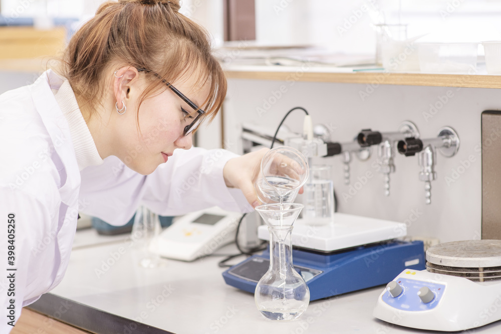 In a chemical laboratory, a girl laboratory assistant pours reagents from a flask into another flask. Assistant in the chemistry laboratory at the university In a chemical laboratory, a girl laboratory assistant pours reagents from a flask into another flask. Assistant in the chemistry laboratory at the university