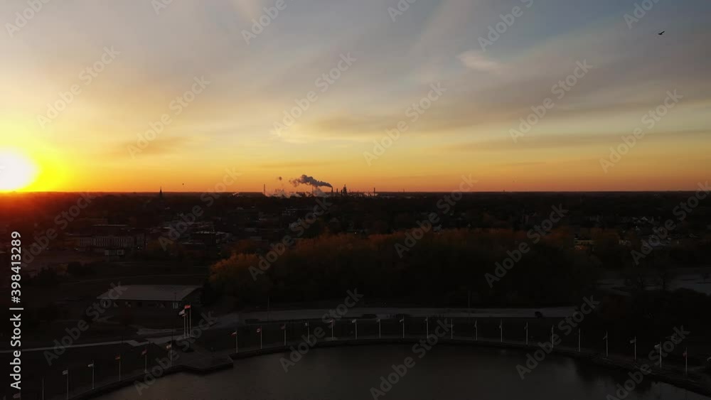 Aerial lowering down towards a waterfront park on the river in Toledo, Ohio as the bright orange sun rises over the horizon lighting up the sky and the smoke spewing from smokestacks on the horizon. Aerial lowering down towards a waterfront park on the river in Toledo, Ohio as the bright orange sun rises over the horizon lighting up the sky and the smoke spewing from smokestacks on the horizon.