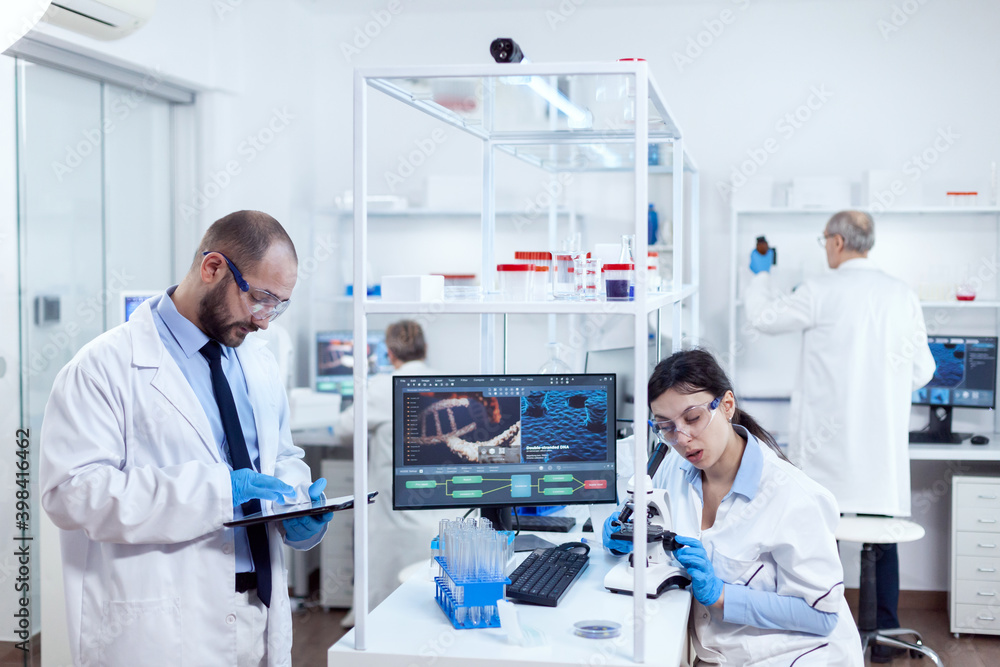 Scientist analysing test results on tablet pc. Team of researchers doing pharmacology engineering in sterile laboratory for healthcare industry with african assistant in the background. Scientist analysing test results on tablet pc. Team of researchers doing pharmacology engineering in sterile laboratory for healthcare industry with african assistant in the background.