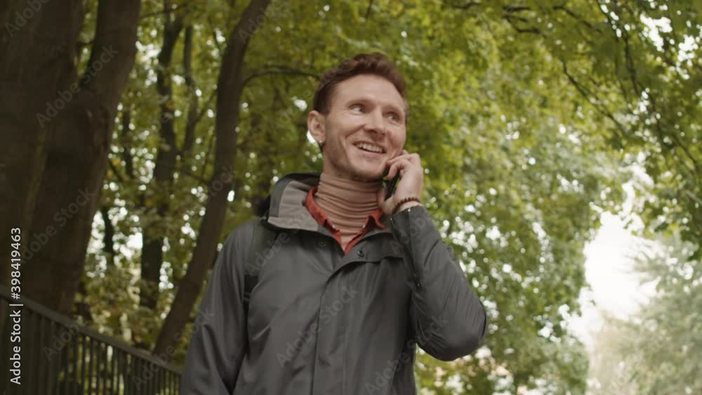 Low angle tracking of middle-aged Caucasian man walking in park, talking on smartphone, waving hand greeting invisible people, smiling Low angle tracking of middle-aged Caucasian man walking in park, talking on smartphone, waving hand greeting invisible people, smiling