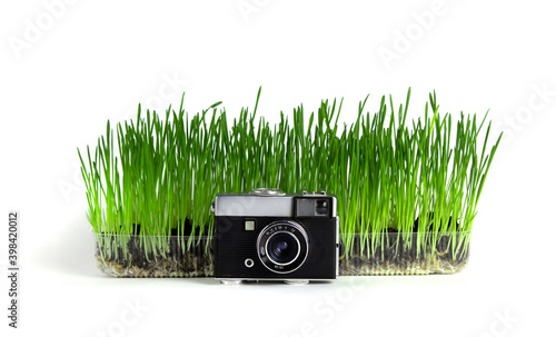 retro camera with small lens stands in front of a container with grass on a white background.