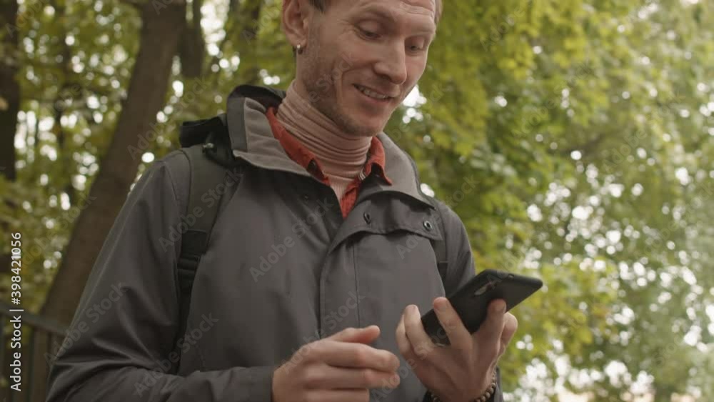 Tilting up of male Caucasian person typing on smartphone, then bringing it to ear and starting talking. Man standing, talking, smiling in public park Tilting up of male Caucasian person typing on smartphone, then bringing it to ear and starting talking. Man standing, talking, smiling in public park