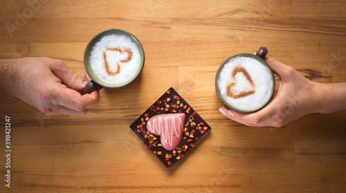 love concept. male and female hands holding identical cups of coffee with milk, with cinnamon hearts, next to them lies a beautiful chocolate heart. Textured beige wooden background. 