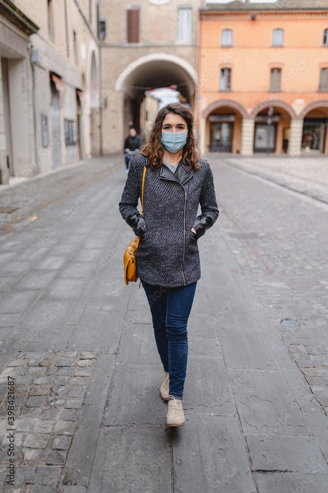 Vertical close-up portrait caucasian young woman walking in a city center during cold winter season. Lady wearing a protective face mask. Pandemic, new normal concept.