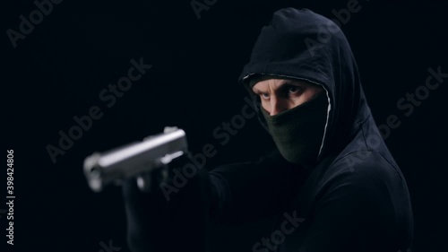 Masked robber in hood aiming with gun in studio. Isolated over black background. Aggressive person threatening someone.