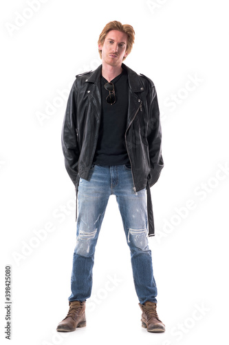 Confident serious young stylish man in jeans and leather jacket posing with hands behind back. Full body length isolated on white background. 