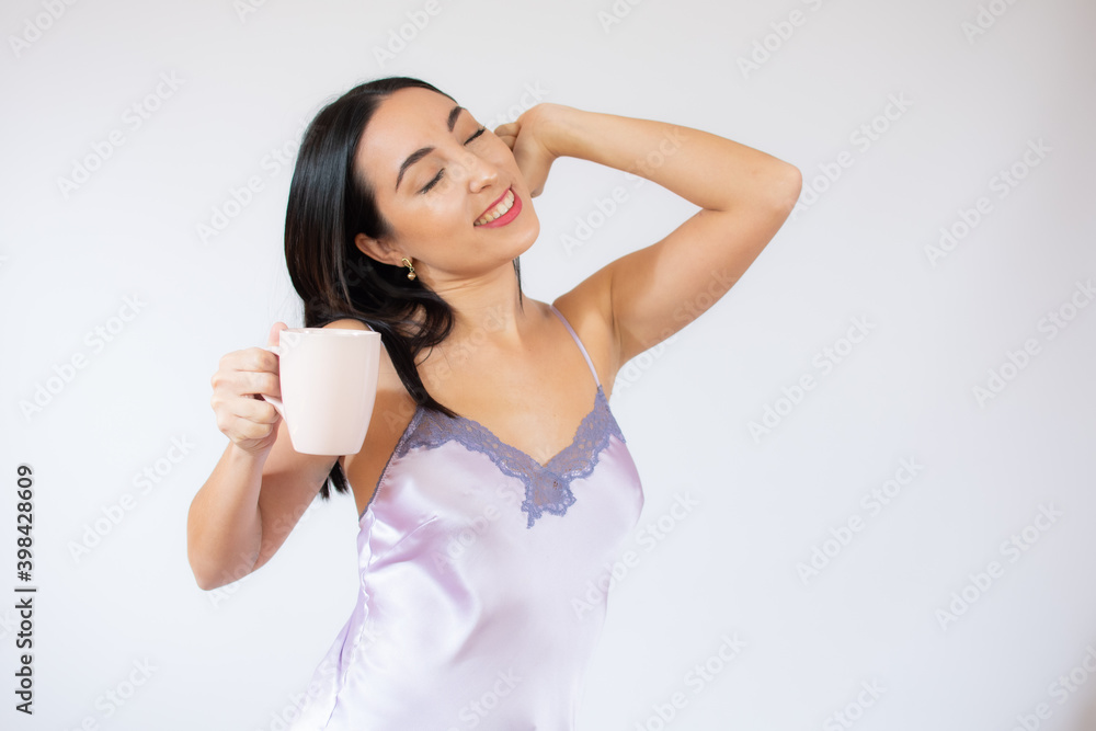 Relaxed joyful woman stretches hands after awakening, rejoices good day, enjoys pleasant sleep, wearing nightgown, has happy expression, poses over white wall Relaxed joyful woman stretches hands after awakening, rejoices good day, enjoys pleasant sleep, wearing nightgown, has happy expression, poses over white wall