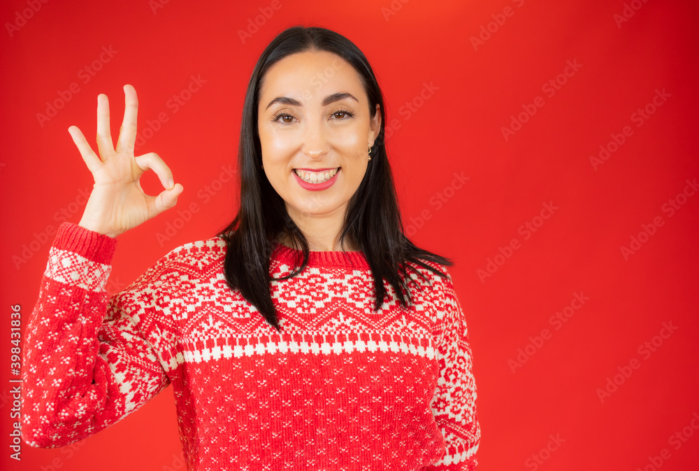 Young beautiful brunette woman wearing casual sweater standing over red background smiling positive doing okay sign with hand and fingers. Successful expression. Young beautiful brunette woman wearing casual sweater standing over red background smiling positive doing okay sign with hand and fingers. Successful expression.