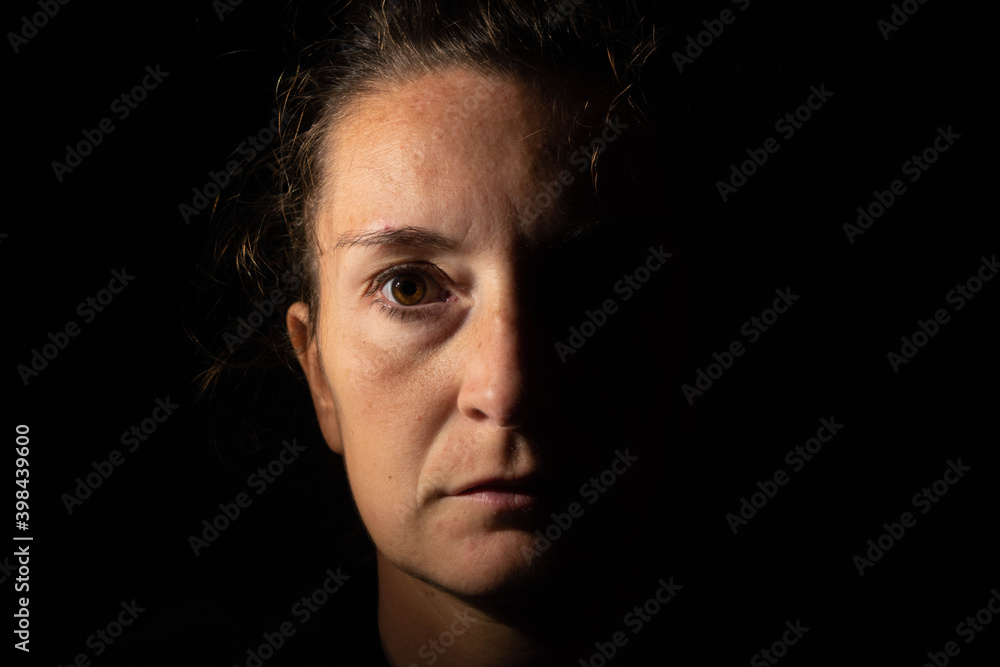 Dark portrait of a serious woman with only half her face lit up on a black background looking sad or in a depression.