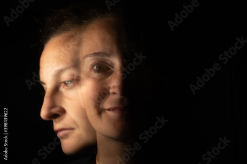 Dark portrait of a smiling woman with only half her face lit up on a black background that unfolds into another blurred face with a sad or depressed gesture.