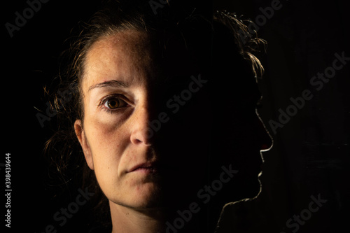 Dark portrait of a serious woman with only half an illuminated face on a black background that is unfolded in the reflection of a mirror in which only the profile of the woman is seen photo