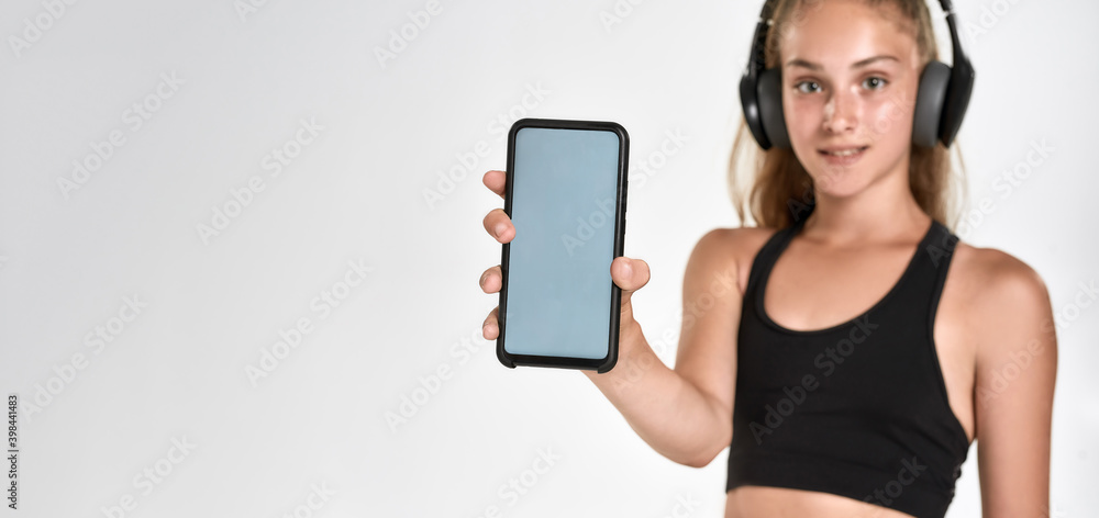 Portrait of cute sportive girl child in headphones, looking at camera, showing smartphone with blank screen, standing isolated over white background