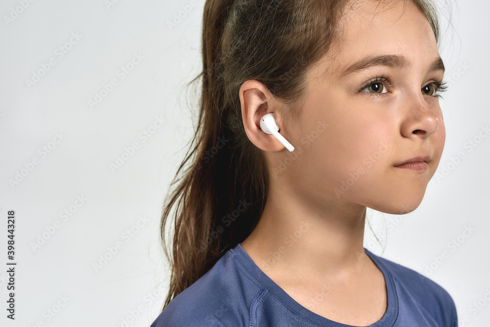 Close up portrait of little sportive girl child wearing earphones, listening to music, looking away while posing isolated over white background Close up portrait of little sportive girl child wearing earphones, listening to music, looking away while posing isolated over white background