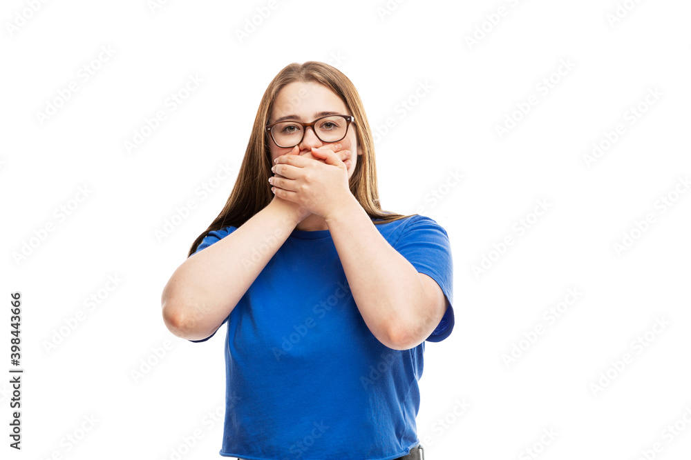 A young brunette girl in glasses in a blue T-shirt covered her mouth with her hands. Isolated on white background. A young brunette girl in glasses in a blue T-shirt covered her mouth with her hands. Isolated on white background.