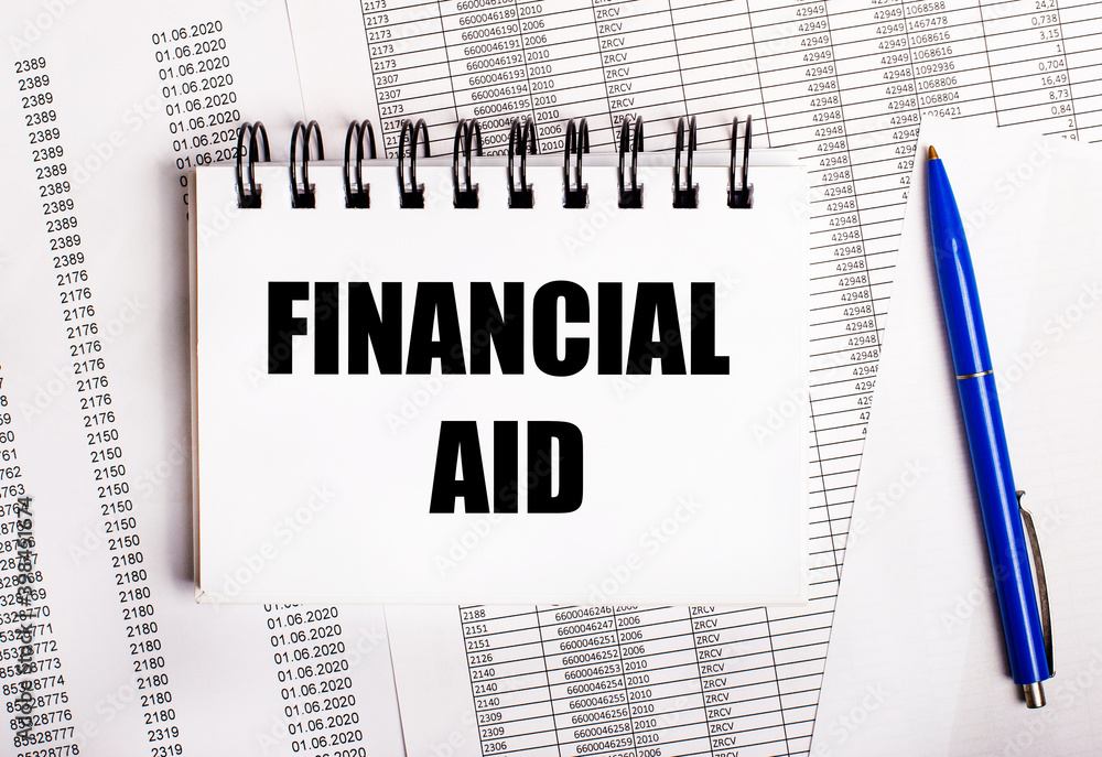 On the table are charts and reports, on which lie a blue pen and a notebook with the word FINANCIAL AID On the table are charts and reports, on which lie a blue pen and a notebook with the word FINANCIAL AID