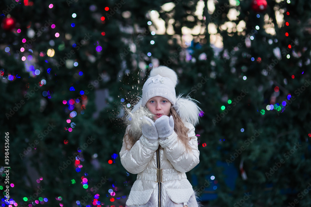 Portrait of a sweet smiling girl in a white knitted cap and white coat standing near decorated Christmas tree on a cold winter day outdoors Portrait of a sweet smiling girl in a white knitted cap and white coat standing near decorated Christmas tree on a cold winter day outdoors