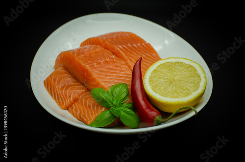 Pieces of Fresh Salmon fillet laying on white plate. isolated on black. Served with chilli pepper, lemon and greens