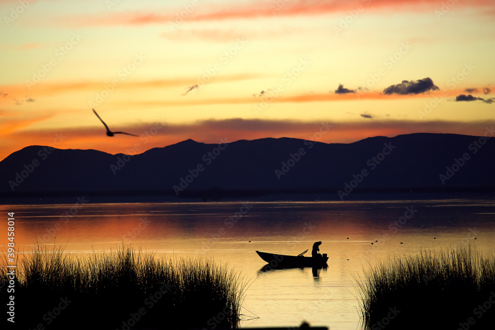 Wonderful sunrise on a lake with a fisherman in his boat and a bird flying towards the mountains in the background