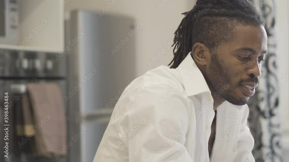 Side view close-up of confident positive African American man sitting in kitchen and talking. Handsome young guy using video chat for remote working indoors. Lifestyle and freelance concept. Side view close-up of confident positive African American man sitting in kitchen and talking. Handsome young guy using video chat for remote working indoors. Lifestyle and freelance concept.