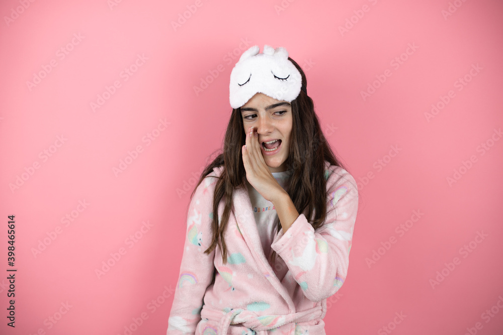 Pretty girl wearing pajamas and sleep mask over pink background with her hand on mouth telling secret rumor, whispering malicious talk conversation Pretty girl wearing pajamas and sleep mask over pink background with her hand on mouth telling secret rumor, whispering malicious talk conversation