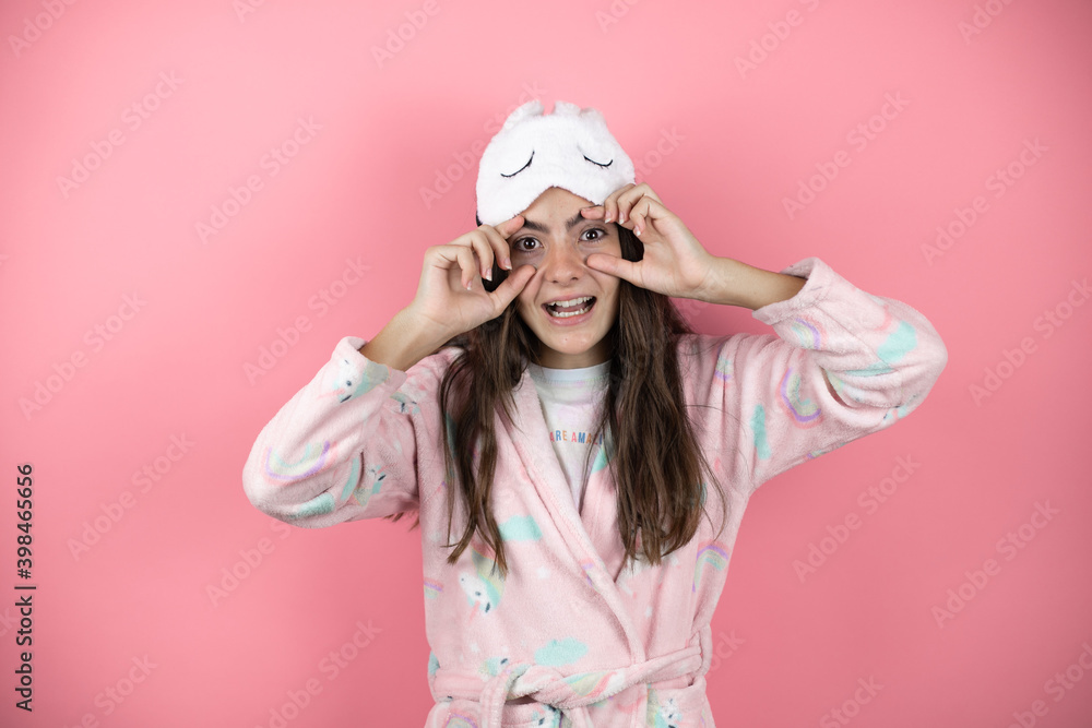 Pretty girl wearing pajamas and sleep mask over pink background Trying to open eyes with fingers, sleepy and tired for morning fatigue Pretty girl wearing pajamas and sleep mask over pink background Trying to open eyes with fingers, sleepy and tired for morning fatigue