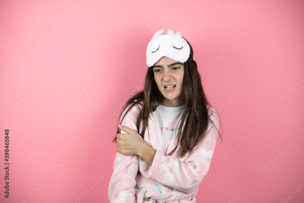 Pretty girl wearing pajamas and sleep mask over pink background with pain on her shoulder and a painful expression Pretty girl wearing pajamas and sleep mask over pink background with pain on her shoulder and a painful expression