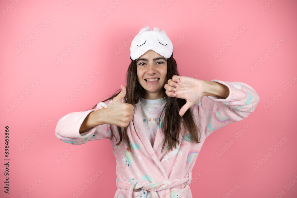 Pretty girl wearing pajamas and sleep mask over pink background Doing thumbs up and down, disagreement and agreement expression. Crazy conflict Pretty girl wearing pajamas and sleep mask over pink background Doing thumbs up and down, disagreement and agreement expression. Crazy conflict