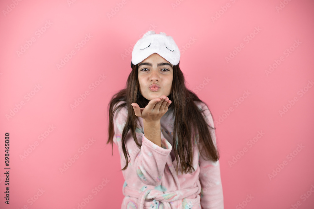 Pretty girl wearing pajamas and sleep mask over pink background looking at the camera blowing a kiss with hand on air being lovely and sexy. Love expression. Pretty girl wearing pajamas and sleep mask over pink background looking at the camera blowing a kiss with hand on air being lovely and sexy. Love expression.