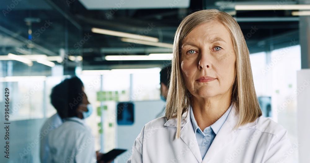 Fototapeta premium Close up portrait of elderly Caucasian woman doctor wearing white coat standing in clinic hall and looking at camera. Mixed-races doctors in masks working on background. Coronavirus concept