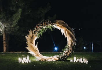 A round, luminous wedding arch, decorated with reeds and glass candlesticks with burning candles and lamps, stands on green grass, against the backdrop of nature in the forest at night.