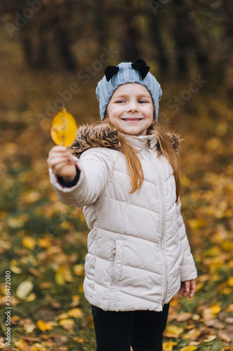 A beautiful little girl, a child in a white jacket of preschool age, stands and poses, holding a yellow wedge-shaped leaf in nature. Autumn portrait, photography.