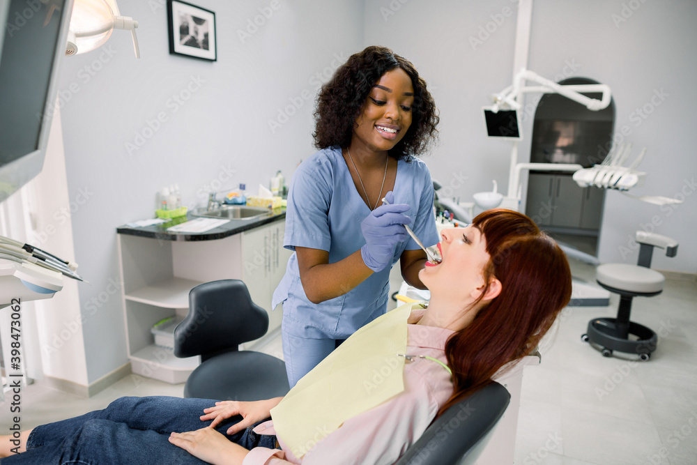 Obraz premium Young female patient at dental office, opening mouth while smiling African woman dentist in uniform and blue latex gloves, checking condition of teeth, using dental mirror. Oral care concept
