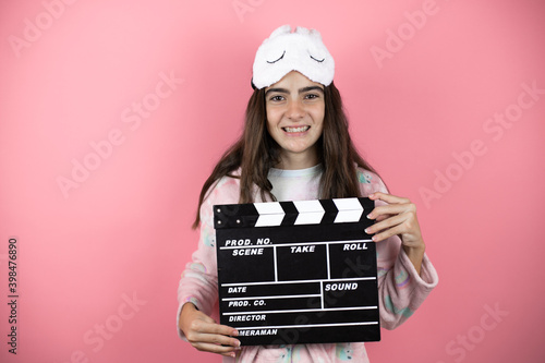 Pretty girl wearing pajamas and sleep mask over pink background holding clapperboard