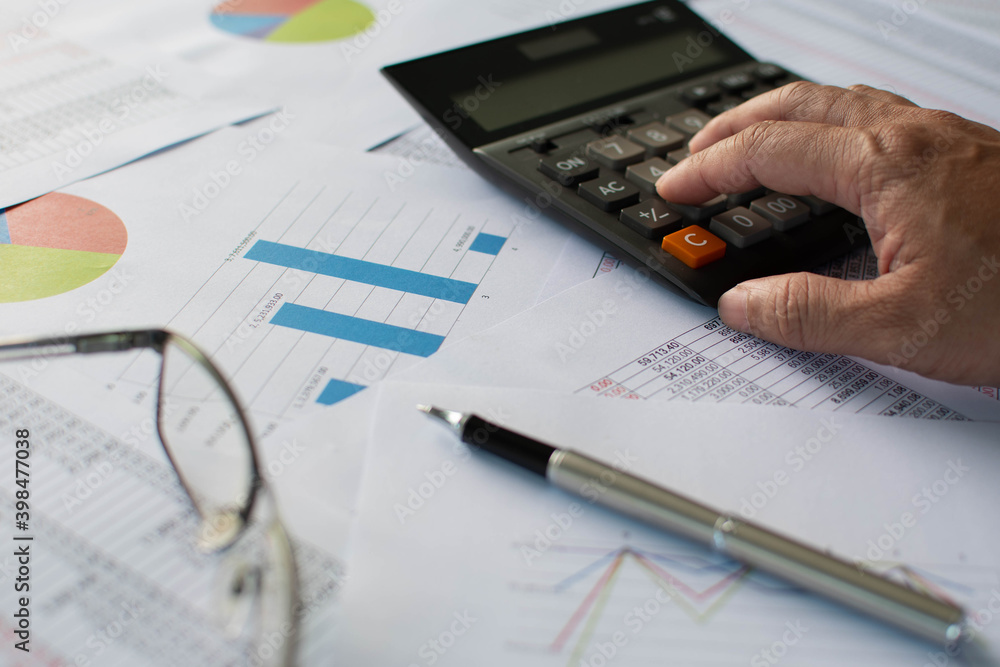 Financial accounting, Closeup, Man's finger pressing a calculator button To calculate financial transaction numbers, A pen, glasses on paper, Graphs showing statistics of financial, Blurred background Financial accounting, Closeup, Man's finger pressing a calculator button To calculate financial transaction numbers, A pen, glasses on paper, Graphs showing statistics of financial, Blurred background
